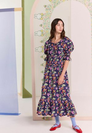 Rochie maxi din bumbac floral, cu mâneci buff și o croială cu volane, având un fundal bleumarin cu flori strălucitoare. Este asortată cu șosete albastre și încălțăminte roșie.