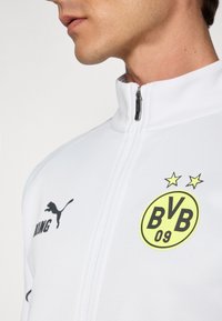Veste blanche zippée avec col montant, arborant un logo jaune de Borussia Dortmund, un logo noir de Puma et un tissu lisse et sportif.