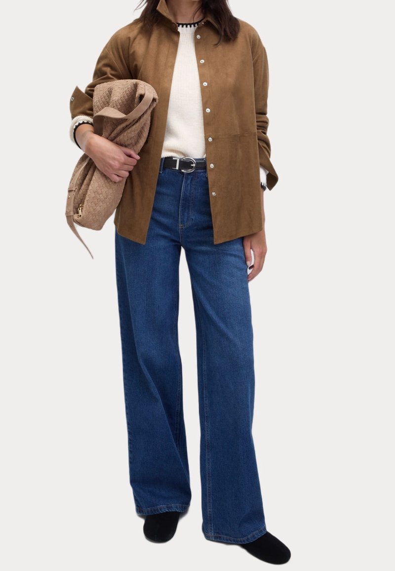 Chemise en suède marron à boutons, pull en tricot blanc, jeans larges bleu, tenant un sac beige, bottes noires, design minimaliste.