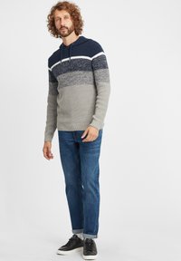 Pleteni kapucar v temno modri, sivi in beli barvi z teksturiranim vzorcem. V kombinaciji z modrimi jeans hlačami in črnimi športniki.