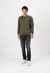 Felpa verde oliva scuro con maniche lunghe e scollatura rotonda. Logo ricamato piccolo sul petto. Abbinata a jeans scuri e sneakers bianche.