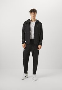 Μαύρο αθλητικό μπουφάν με κουκούλα και τσέπες με φερμουάρ, συνδυασμένο με ταιριαστά παντελόνια. Λευκά λογότυπα και ελαφρύ, υφασμένο ύφασμα.