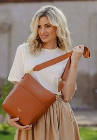Hnědá kožená crossbody taška se strukturovaným tvarem, zlatými kovovými detaily a nastavitelným popruhem, kterou drží osoba oblečená v bílém topu.