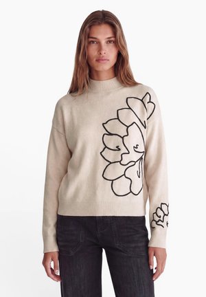 Vrouw die een beige trui draagt met grote zwarte bloemcontouren aan de voorkant en mouw, gecombineerd met zwarte jeans, staand tegen een witte achtergrond.