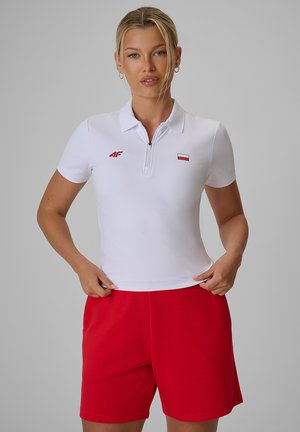 Kobieta o blond włosach, ubrana w białą koszulkę polo z krótkim rękawem i czerwone szorty, stojąca na tle jednolitego szarego tła.