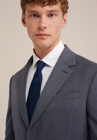 Grijs pak met een gestructureerde afwerking, voorzien van een notch-kraag, hoge kraag en een marineblauwe stropdas. Het overhemd is wit, wat zorgt voor een formele uitstraling.