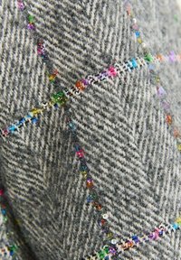 Detailní záběr na šedou látku s herringbonovým vzorem, na které jsou barevné korálky a flitry umístěné podél našitých diamantových vzorů.