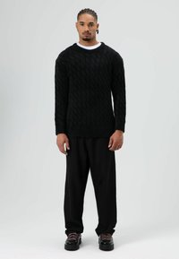 Zwarte kabelgebreide trui in een losse pasvorm, gecombineerd met losse zwarte broek en stevige zwarte schoenen met roze veters, tegen een lichte achtergrond.