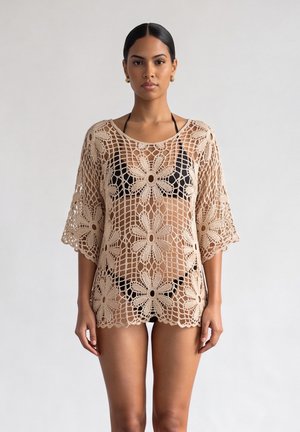 Vrouw draagt een beige gehaakte cover-up met bloempatronen over een zwarte bikini, rechtopstaand tegen een effen lichte achtergrond.
