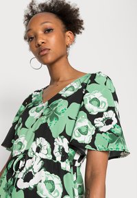 Vestido floral em preto com grandes padrões de flores verdes e brancas, decote em V, mangas curtas e cintura ajustável com cordão.