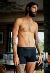 Sorte bomulds boxer briefs med en DOVRE logo talje, tætsiddende design og detaljerede arm tatoveringer på en overkropsbar mand i en hyggelig setting.