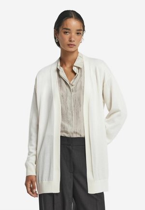 Hvid cardigan lavet af blødt stof, designet med åben front og lange ærmer, parret med en lys stribet bluse og mørke bukser.