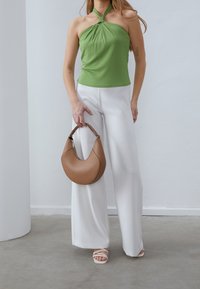 Žena v zeleném topu na ramínka a bílých širokých kalhotách držící hnědou kabelku ve tvaru půlměsíce a obutá v bílých sandálech na podpatku.