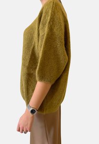 Maglione a maglia verde oliva con scollo rotondo, maniche corte a sbuffo e una texture morbida. Indossato sopra pantaloni crema, che rivelano un orologio.