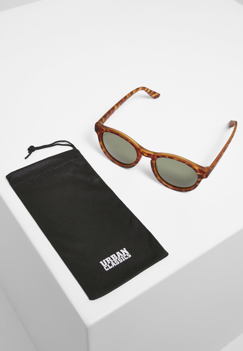 Sunglasses iš tortoškės su žaliais lęšiais, apvalių rėmų. Pridedama juoda raišteliu užsegama dėklas su "URBAN CLASSICS" logotipu.