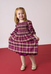 Rochie cu carouri mov, cu mâneci lungi, cu bust smocking și fustă evazată. Asociată cu colanți deschiși și pantofi strălucitori, pe un fundal roșu.