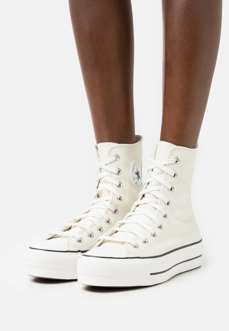 Personne à la peau foncée portant des baskets blanches montantes Converse Chuck Taylor All Star avec des rayures noires sur fond blanc.