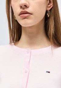 Κοντινό πλάνο ενός ατόμου που φοράει ροζ ανοιχτό χρώμα πουκάμισο με κουμπιά, με μικρό κεντητό λογότυπο και χρυσούς κρίκους για σκουλαρίκια.