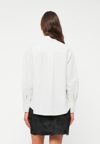 Camicia bianca a maniche lunghe con polsini abbottonati, caratterizzata da leggere pieghe e un orlo curvo, abbinata a una gonna scura ricamata con fiori.
