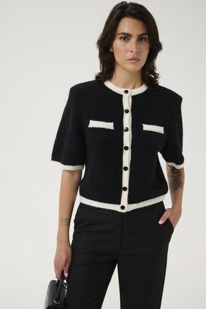 Cardigan noir en tricot à manches courtes, avec des bordures blanches, deux poches poitrine et des boutons noirs. Assorti avec un pantalon noir ajusté.