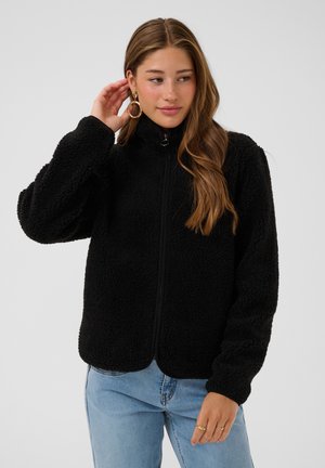 Young woman with long hair wearing a black zip-up fleece jacket and light blue jeans adjusting her large hoop earring.
