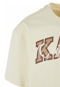 Cream cotton t-shirt with short sleeves. Features a large, brown graphic text design on the front. Soft texture and relaxed fit.