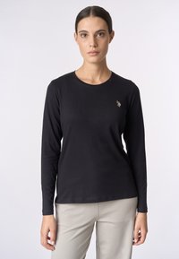 Donna che indossa una maglietta nera a maniche lunghe con un piccolo logo di un giocatore di polo sul petto e pantaloni beige chiari, in piedi contro uno sfondo semplice.