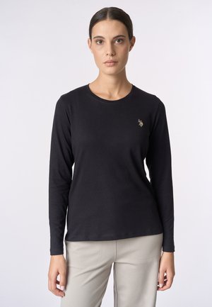 Donna che indossa una maglietta nera a maniche lunghe con un piccolo logo di un giocatore di polo sul petto e pantaloni beige chiari, in piedi contro uno sfondo semplice.
