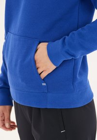 Blå sweatshirt med en frontlomme, ribbede manchetter og kant. Har en glat tekstur og sidder over sorte bukser.