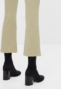 Jeans beige acampanados con dobladillo sin terminar, combinados con botas de tobillo negras con tacón grueso y material liso y ajustado.