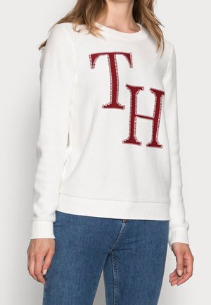 Femme portant un pull en maille blanc avec de grandes lettres rouges "TH" sur le devant et un jean bleu, debout devant un fond uni.