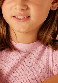 Roze gebreide top met een textuur van diamantpatronen, ronde halslijn en korte mouwen. Klein hartvormig oorbellen zichtbaar.
