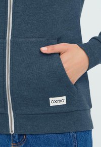 Tumši zili kapuci ar rāvējslēdzēju, izgatavots no gludas auduma, ar priekšējo kabatu un "oxmo" markas etiķeti, kas izšūta apakšējā kreisajā stūrī.