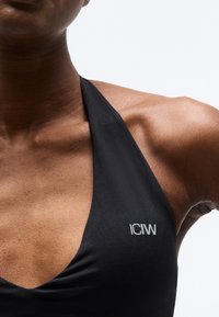 Κοντινό πλάνο ενός ατόμου που φοράει μαύρο τοπ με λαιμόκοψη halter και λογότυπο "ICIW" στο στήθος, σε λευκό απλό φόντο.