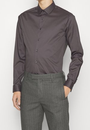 Hombre con una camisa de botones de color púrpura oscuro metida en un pantalón de vestir gris texturizado, con una mano en el bolsillo y fondo neutro.