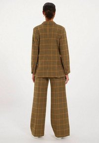 Costume à carreaux comprenant un long blazer ajusté et un pantalon large, fabriqué en tissu texturé dans des tons de jaune, marron et orange.