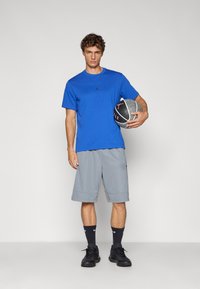 Tricou albastru din bumbac cu un logo negru, asortat cu pantaloni scurți gri și pantofi sport negri, ținând o minge de baschet cu un model.