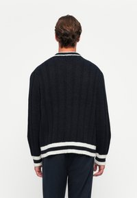 Homme de dos, vêtu d'un pull à côtes sombres avec des poignets et un ourlet à rayures blanches, devant un fond uni.
