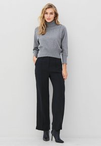 Pilkas turtleneck megztinis su dekoratyviniais kaiščiais peties srityje, derinamas su juodomis aukšto liemens kelnėmis ir smailiais juodais auliniais batais.