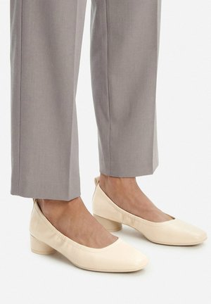 Füße, die beige, flache, geschlossene Schuhe tragen, kombiniert mit hellgrauen, gerade geschnittenen Hosen, die oberhalb der Knöchel enden.