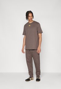 T-shirt en coton marron avec un logo jaune, associé à un pantalon de survêtement marron assorti présentant des motifs texturés subtils, et des chaussures noires.
