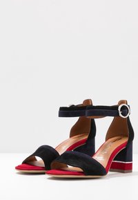 Eleganckie, granatowe zamszowe sandały na obcasie z paskiem na kostce i srebrną klamrą. Czerwone i przezroczyste akcenty na blokowym obcasie. Model z odkrytymi palcami.