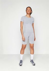 Sieviete stāv gaismas pelēkā sporta tērpā ar šortiem, T-kreklu, baltām vidēja garuma zeķēm un pelēkiem skriešanas apaviem uz vienkārša gaiša fona.