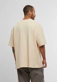 Tricou oversize bej cu un design texturat, înfășurat. Mâneci scurte și decolteu rotund. Vedere din spate care dezvăluie o croială largă și tiv drept.