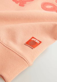 Roza pulover z teksturirano tkanino, z izvezenim vzorcem in rdečim Superdry logotipom, ki je našit na rokavu.
