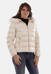 Giacca imbottita color crema con cappuccio in pelliccia sintetica, chiusura a zip e due tasche laterali, caratterizzata da un motivo trapuntato e dettagli lucidi.