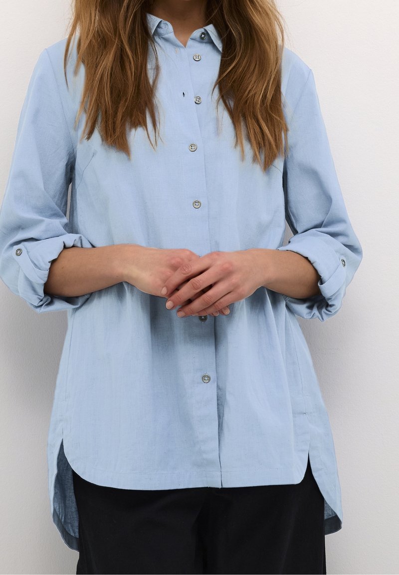 Camicia button-up azzurro chiaro con colletto, maniche lunghe arrotolate ai polsini e spacchi laterali per facilitare i movimenti. Tessuto dalla consistenza liscia.