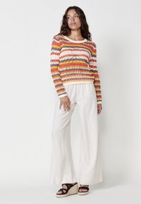 Suéter de punto con rayas coloridas en naranja, crema y gris; combinado con pantalones de lino claros y fluidos, y sandalias de esparto negras con tiras.