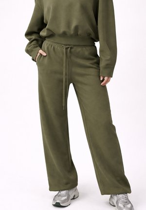 Persoon draagt olijfgroene wijde sweatpants met een trekkoord in de taille en een bijpassende sweater, gecombineerd met grijze en witte sneakers.