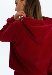 Bordowa, krótka bluza z kapturem o strukturalnej fakturze, z sznurkiem przy kapturze i rękawami raglanowymi. Widok z tyłu pokazuje szczegóły kaptura oraz luźny krój.